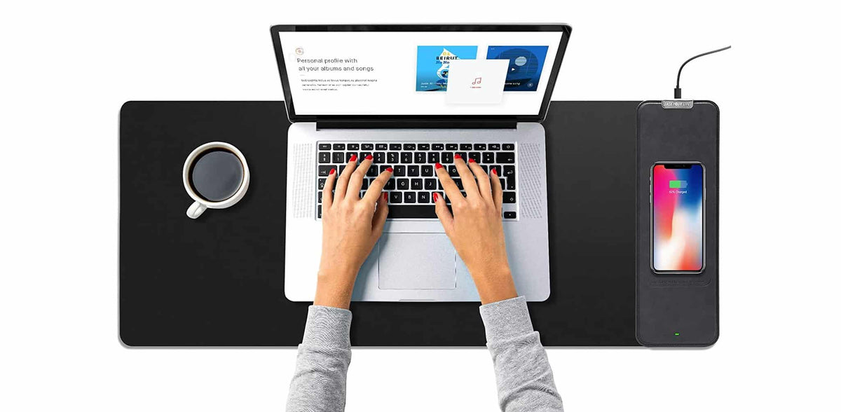 Black 4-in-1 laptop sleeve unfolded as a mouse pad and desk mat with built-in wireless charging pad charging a phone beside a laptop